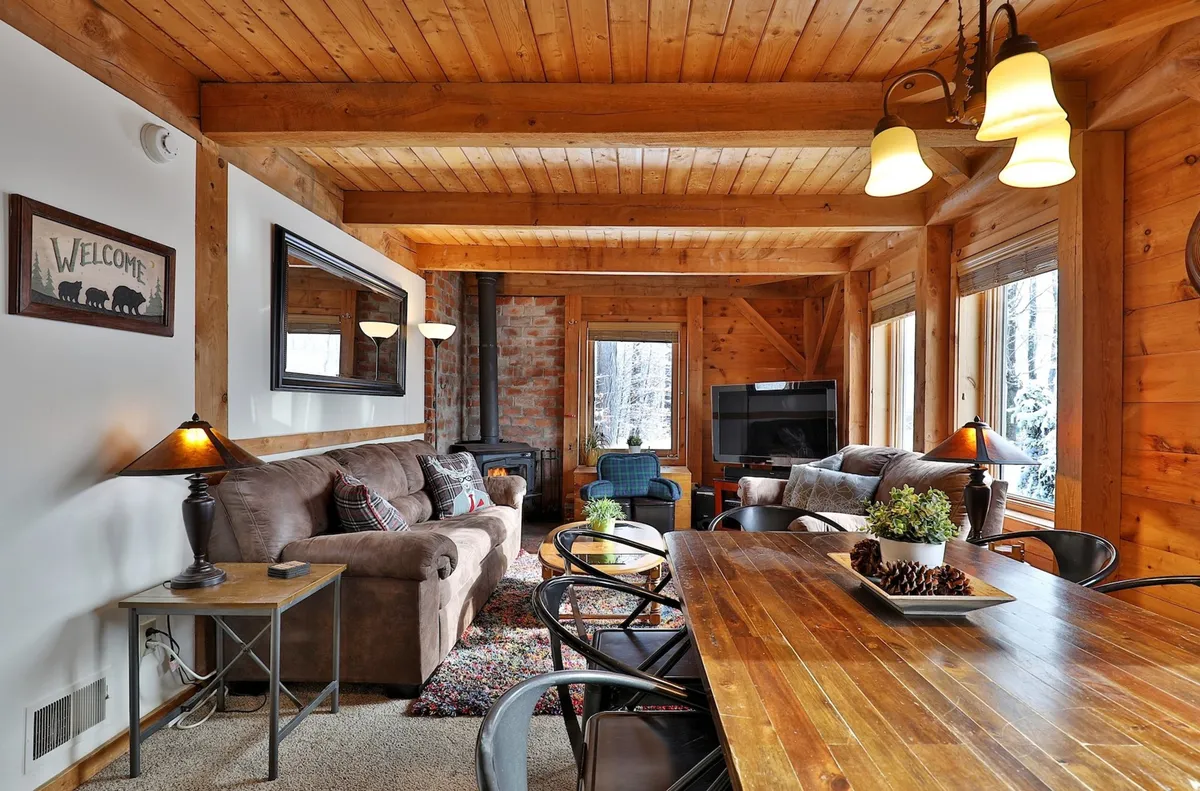 A spacious living area with a wooden dining table, comfortable sofa, stone fireplace, and large windows revealing snowy landscapes outside. The welcome sign adds a personal touch, while the layout demonstrates ample space for relaxation and group activities.