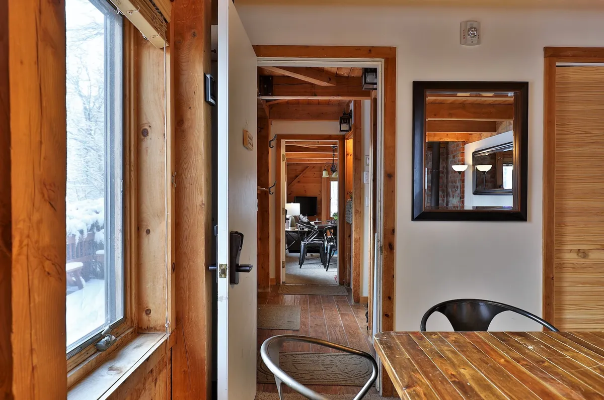 A cozy dining and living space with exposed wooden beams, a generous dining table, large windows framing scenic outdoor views, and open doorways hinting at easy access to the outdoors.