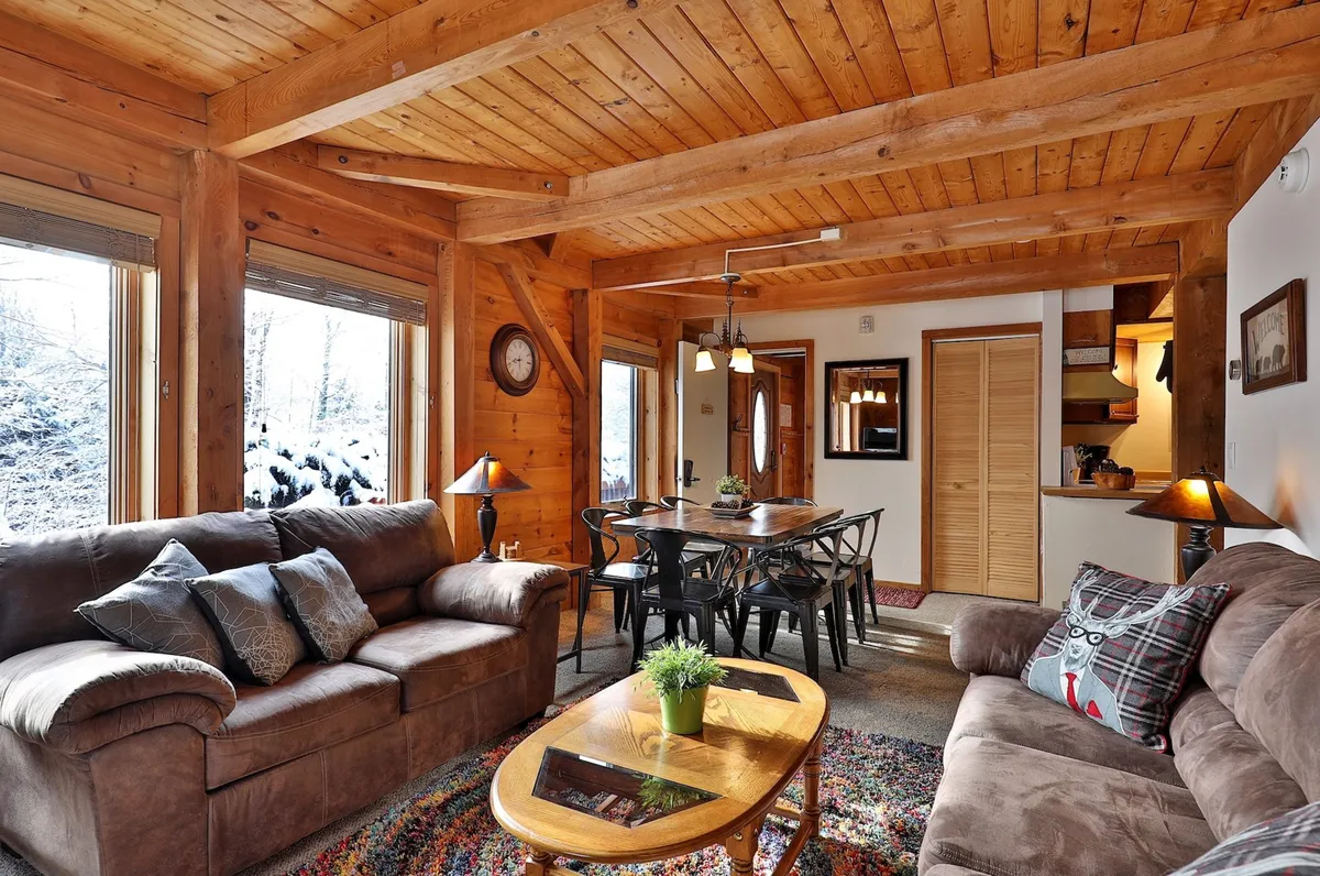 A spacious living area with prominent wooden beams, large windows that let in natural light, comfortable sofas, and a dining table, all arranged in a way that feels open and connected to the outdoors.