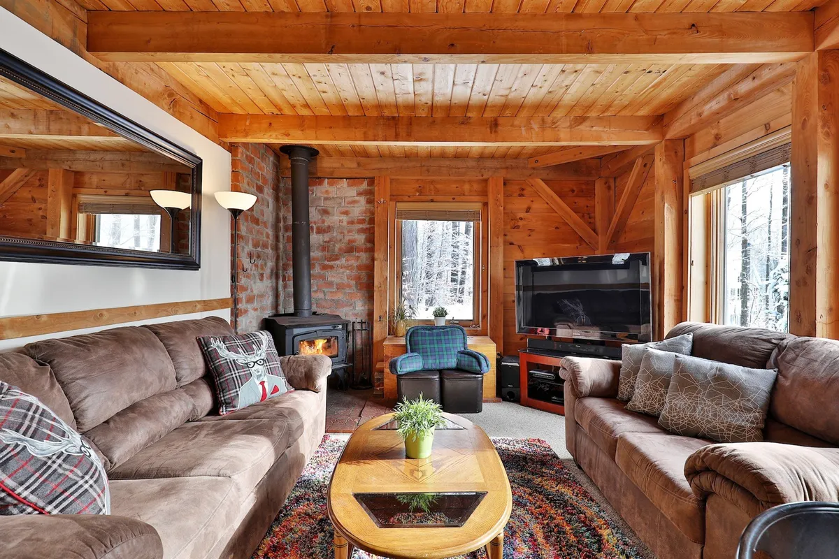A cozy living room featuring exposed wooden beams, a brick stove, large windows letting in natural light, and plush sofas arranged for conversation or lounging. There’s a coffee table for games or drinks, creating a perfect spot for families and friends to unwind.