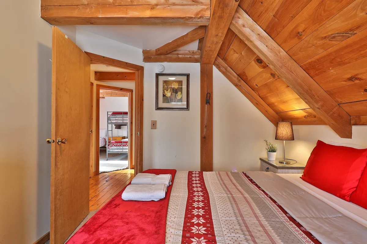A serene queen bedroom featuring a striking red bedspread, rustic wooden beams, framed artwork, and a softly glowing table lamp. Natural light filters through the window, creating an inviting atmosphere ideal for couples or those seeking a quiet retreat.