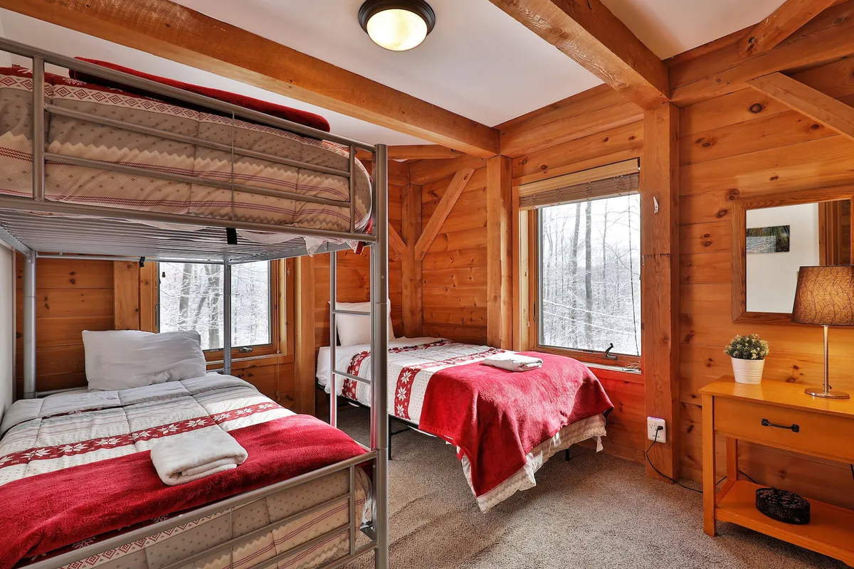 A spacious bedroom featuring a bunk bed and a single bed, both with cozy red blankets. Warm wooden beams and large windows bring in ample natural light, and a table lamp sits next to the beds for bedtime reading. The room feels both comfortable and flexible, suitable for families or groups.