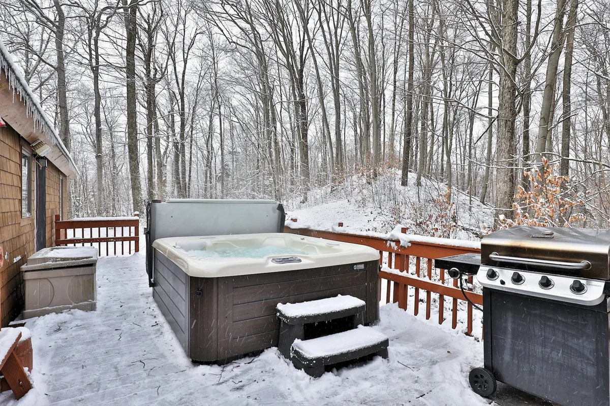 A cozy outdoor scene showing a hot tub and grill set up on a wooden deck, with a backdrop of surrounding trees and a deck railing. The atmosphere feels private and inviting, ideal for both winter and summer seasons. The setup captures the essence of stargazing from the hot tub and the potential for barbecuing no matter the weather.