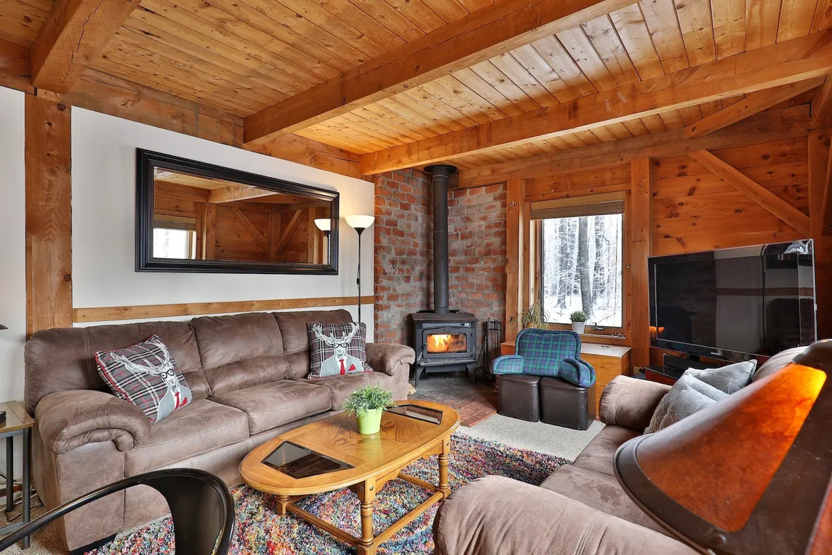 This image showcases a welcoming living room anchored by a stone fireplace, with a comfortable sofa, plaid armchair, and wooden table. Large windows allow natural light, and the overall setting encourages family relaxation, cozy gatherings, and watching TV together.