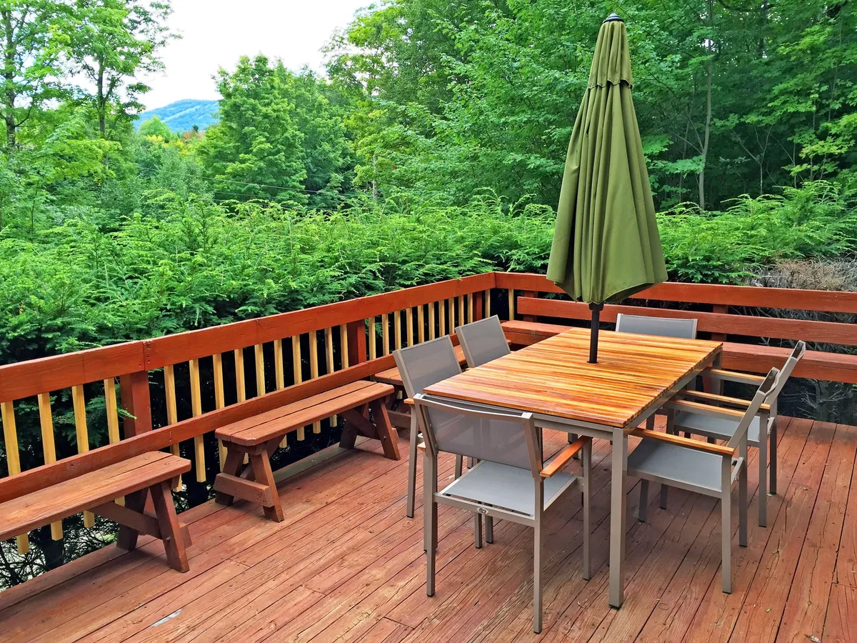 The photo captures a wooden outdoor table with benches and a green umbrella, set amidst beautiful trees and foliage. The scene evokes a sense of retreat and relaxation in nature, perfect for group gatherings or quiet moments.