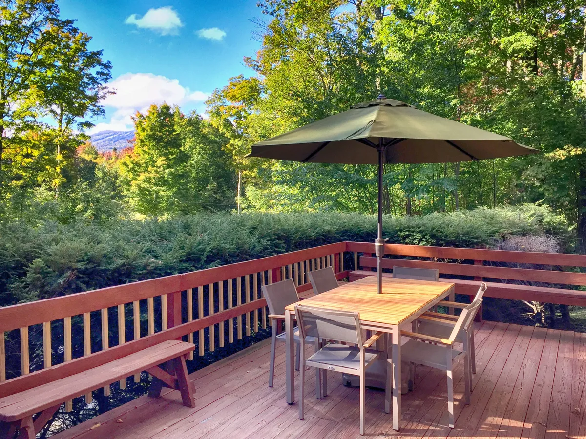 This exterior image features a dining table with chairs and a patio umbrella, set against lush surrounding greenery. The scene suggests a peaceful outdoor space for dining and socializing, with natural light and a tranquil atmosphere.