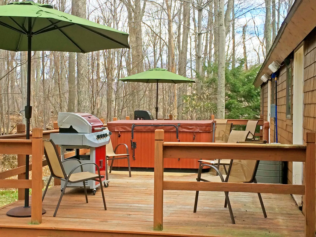 A vibrant outdoor patio area equipped with a grill, hot tub, patio furniture, and umbrellas. The space is set for socializing and leisure, with the inviting hot tub and grill suggesting both relaxation and outdoor gatherings.