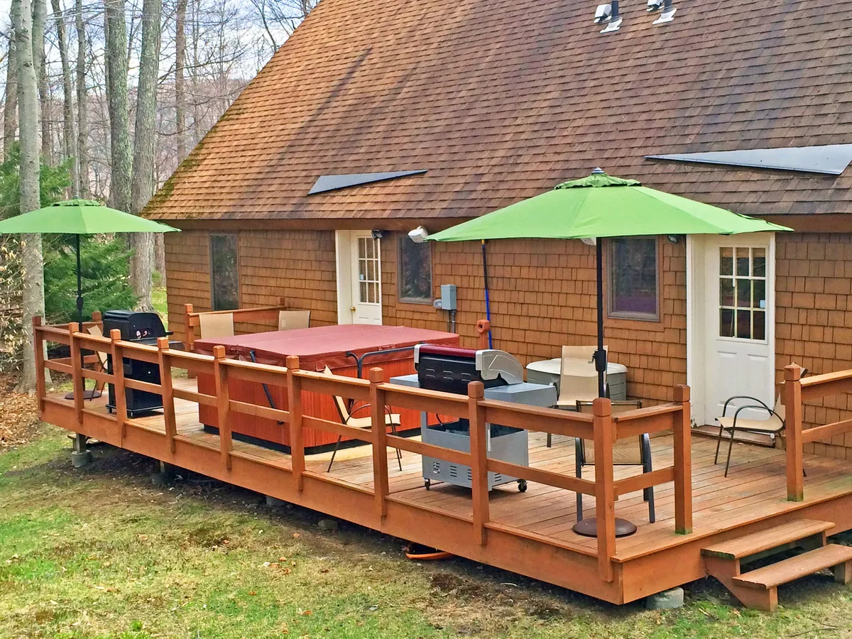 An expansive deck scene featuring a hot tub, green umbrellas, a grill, and comfortable outdoor seating. The setup is perfect for a variety of activities, from barbecuing to lounging and soaking in the tub. The space is bright and welcoming, suggesting both daytime and evening enjoyment.