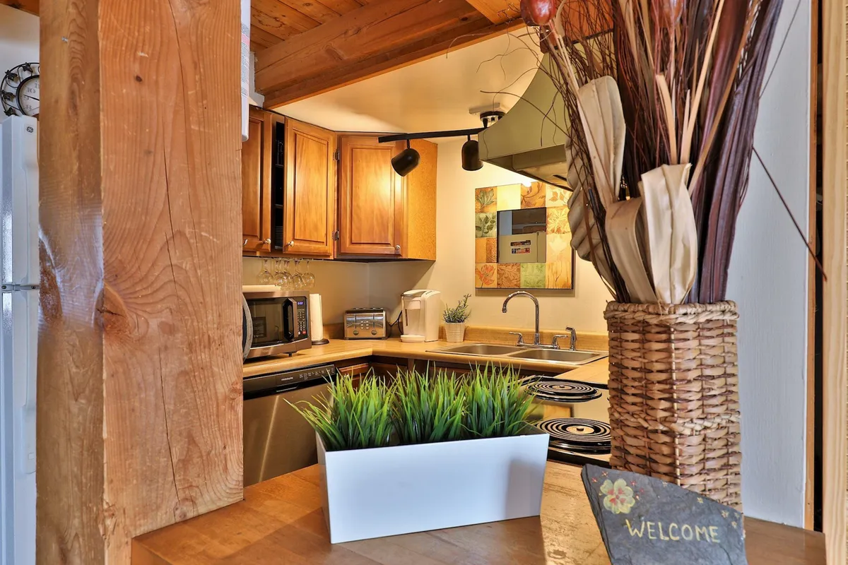 A warm, inviting kitchen scene with visible kitchen appliances, wooden cabinetry, a decorative plant, and a welcome sign. The countertop is ideal for meal prep, and the space suggests the presence of multiple refrigerators and ovens.